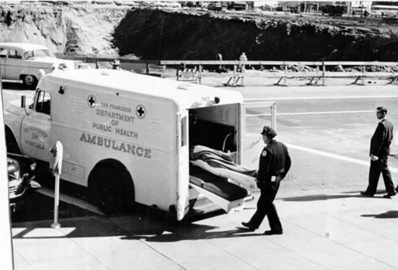 San Francisco Department of Public Health ambulance with its rear doors open and a gurney being loaded, attended by a uniformed officer